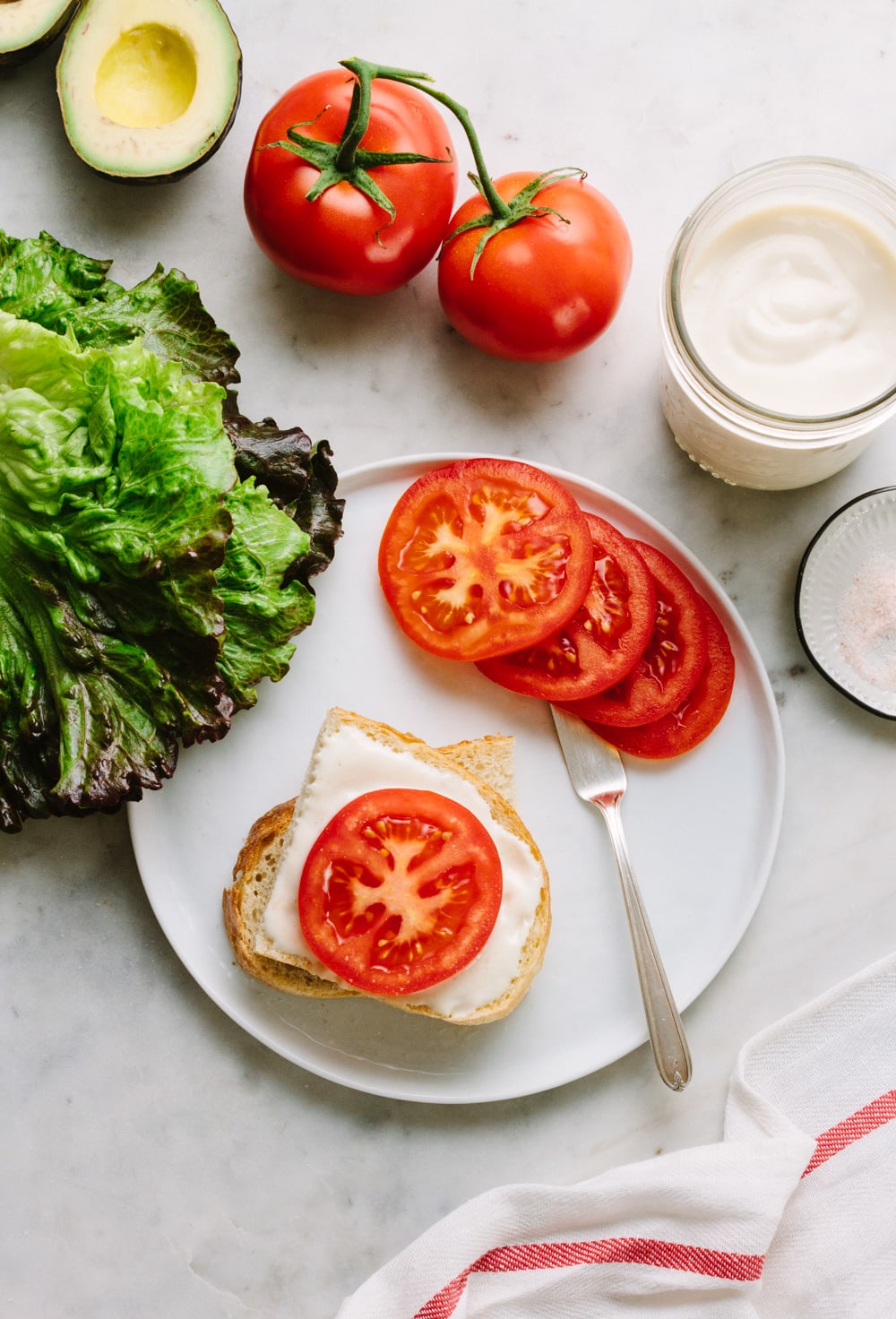 creamy vegan mayonnaise spread on a slice of artisan bread and topped with a tomato on a white plate surrounded by leafy greens
