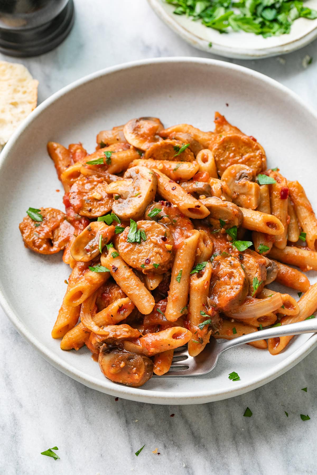 side angle view of plated serving of vegan sausage pasta with mushrooms in a shallow bowl with items surrounding.