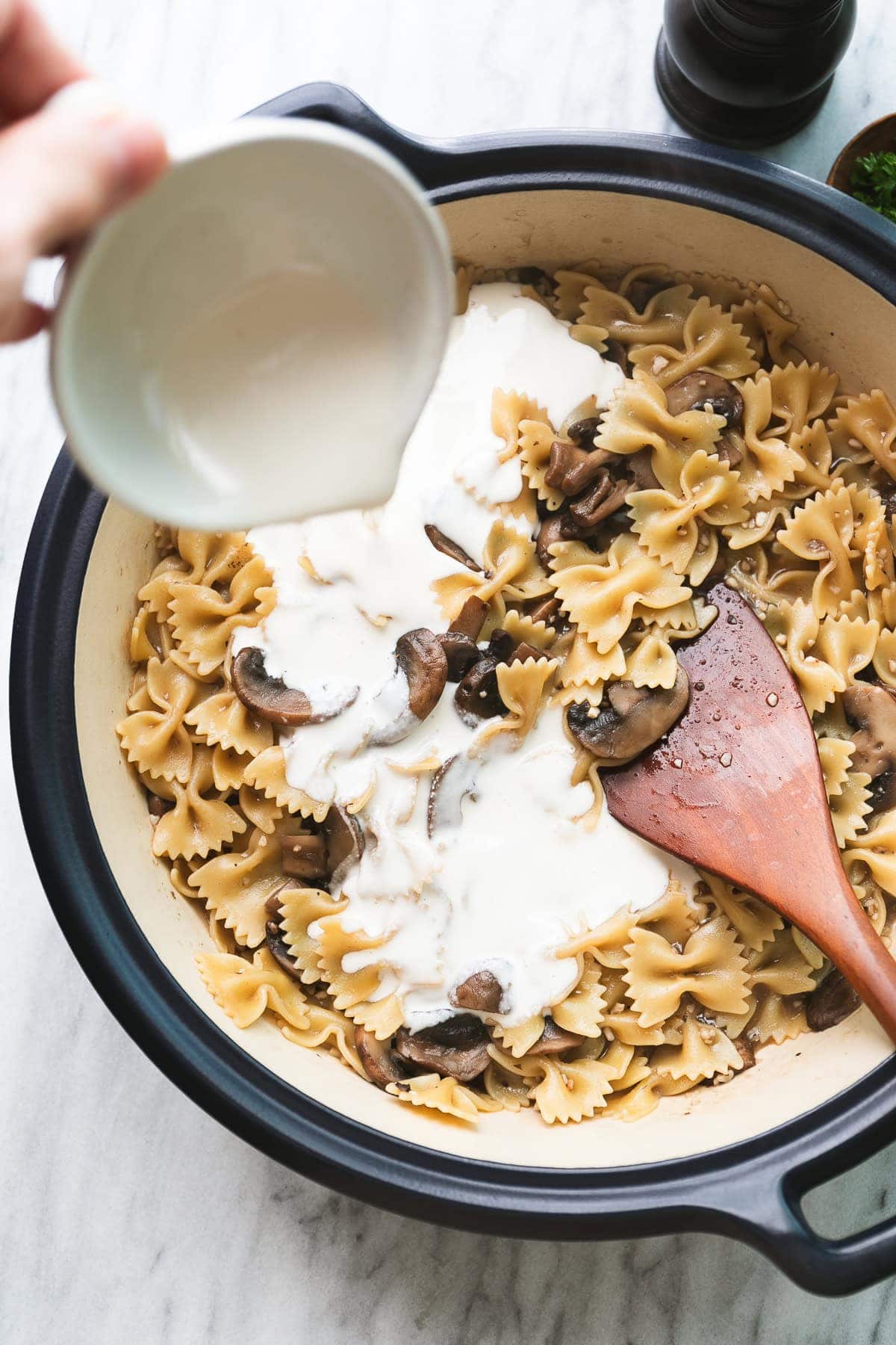 top down view of vegan cream being added to mushroom pasta in a pot with wooden spoon.
