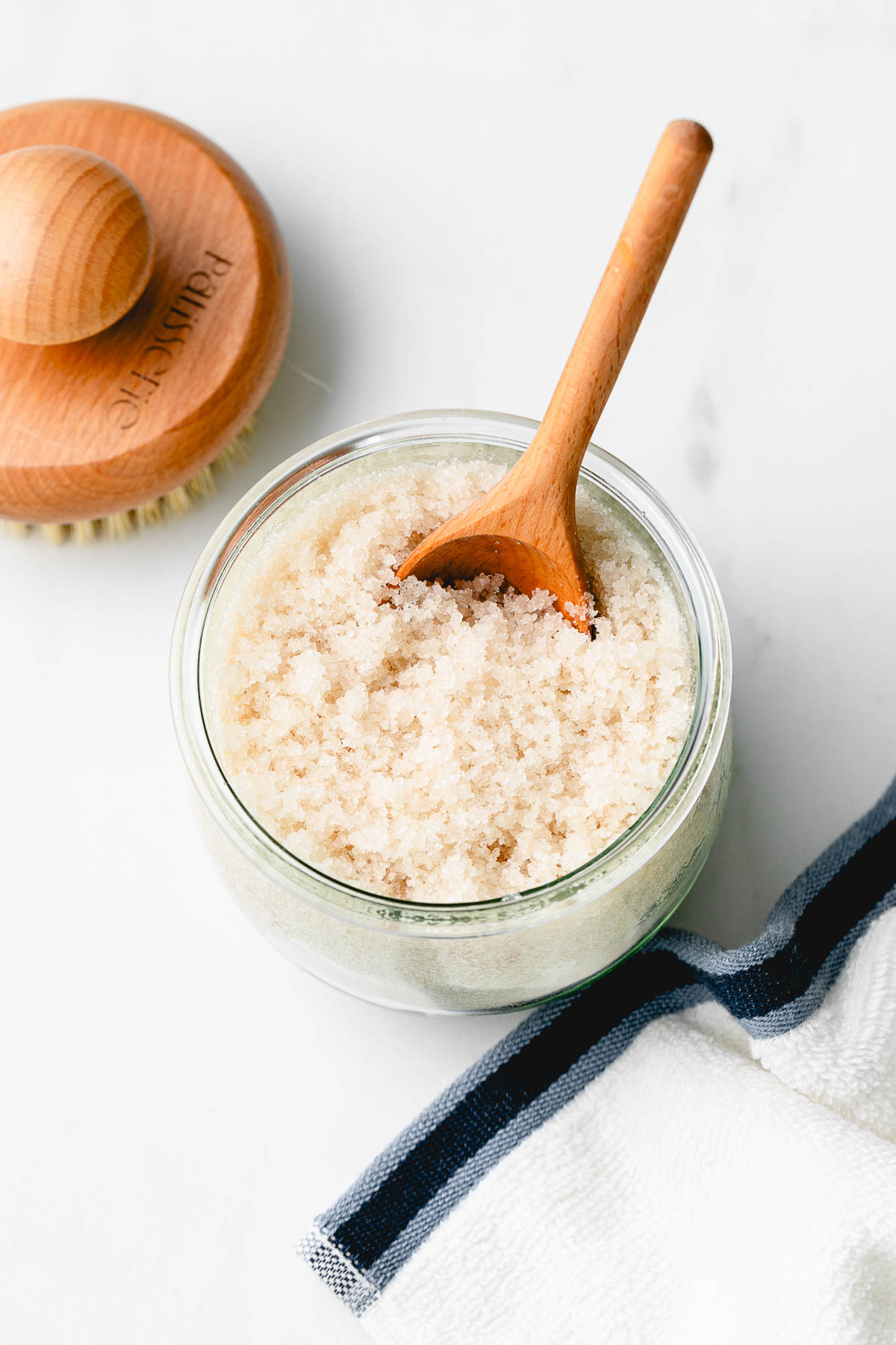 side angle view of sugar scrub in a jar with wooden spoon and items surrounding.