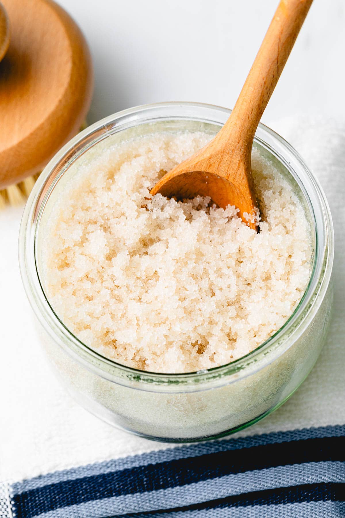 side angle view of sugar scrub in a jar with wooden spoon and items surrounding.