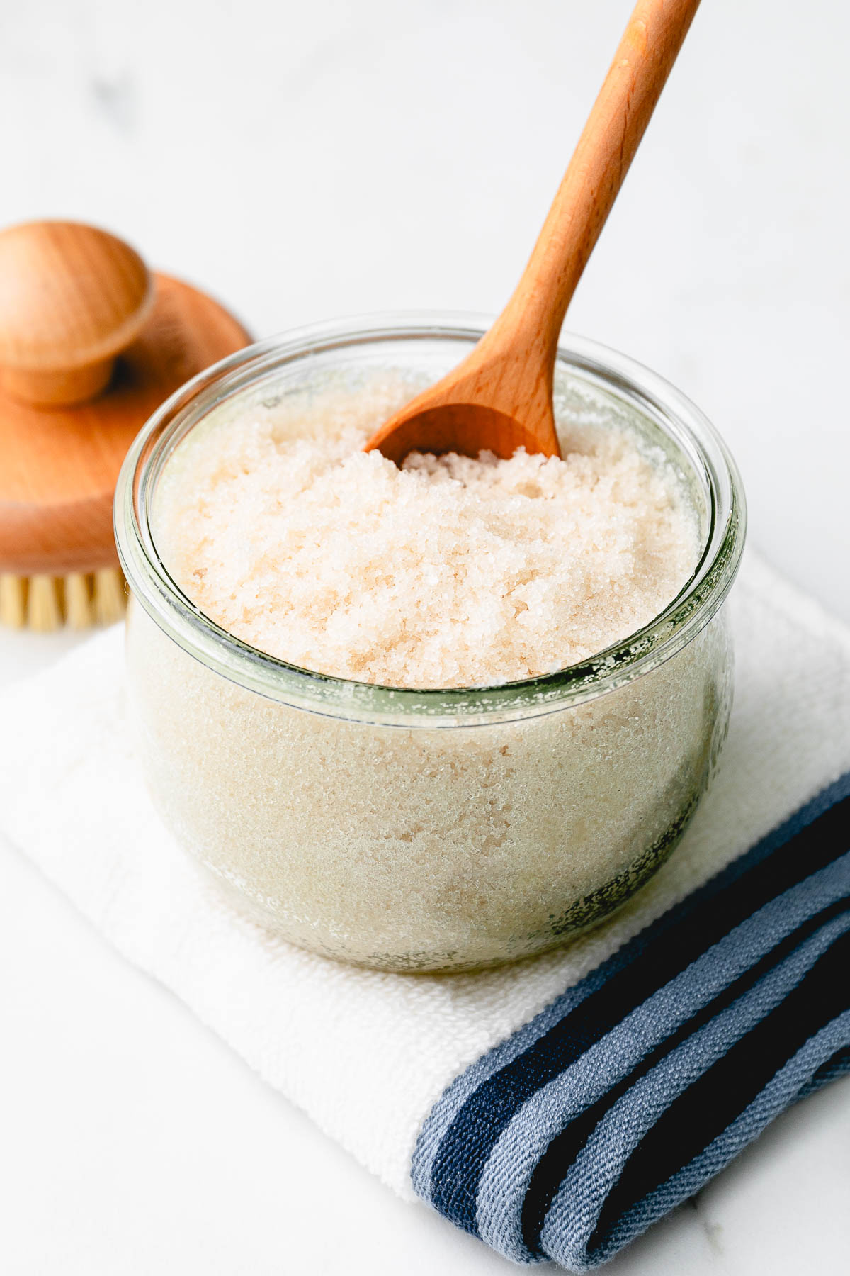 side angle view of sugar scrub in a jar with wooden spoon and items surrounding.