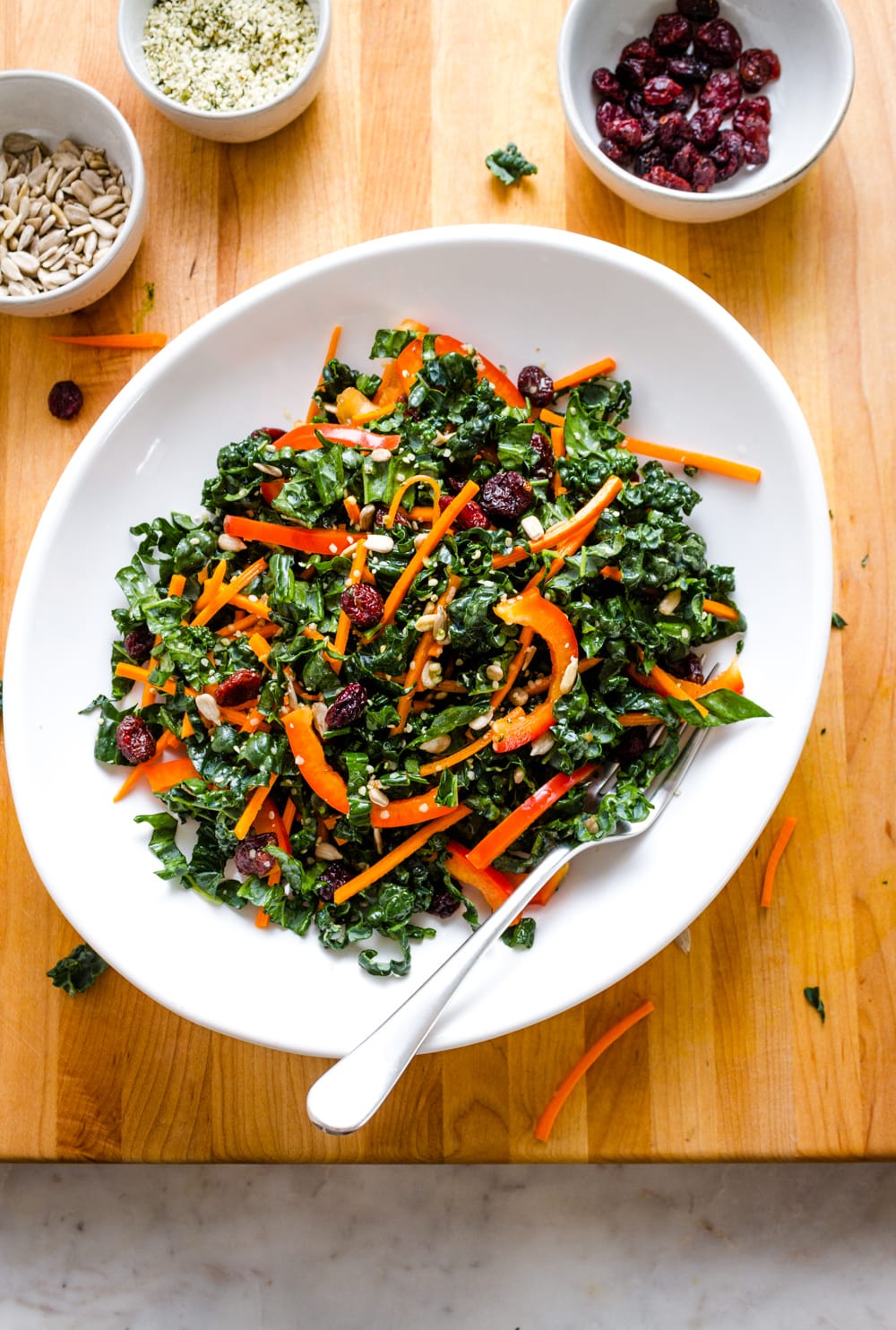 top down view of kale salad with cranberries in a white bowl with fork, sitting on a wooden cutting board.