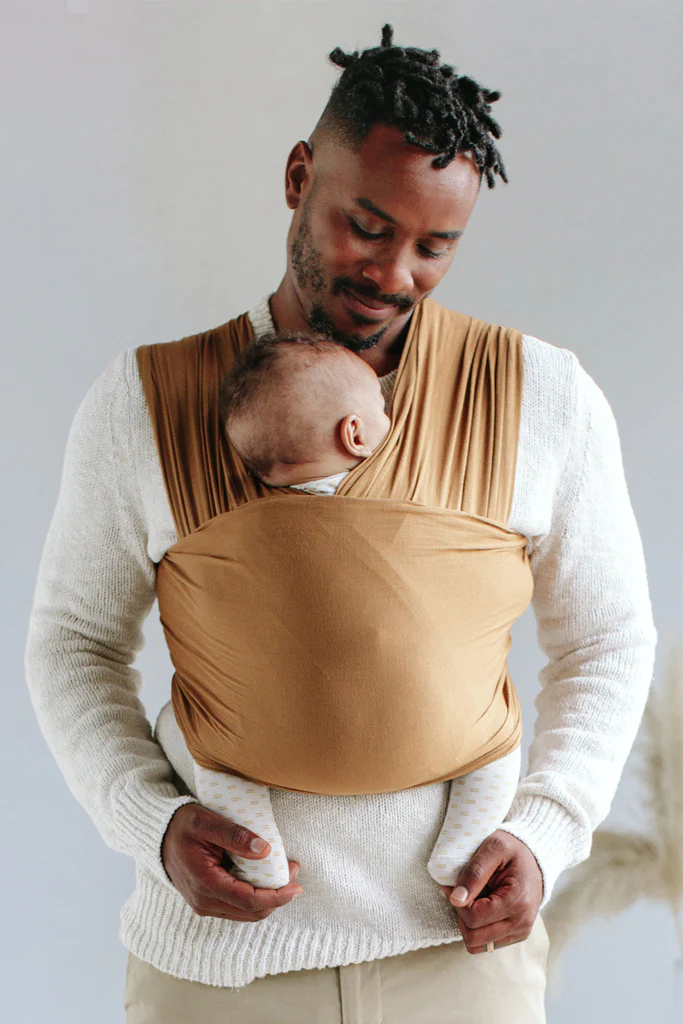 A man holds a baby, who is wrapped to his chest with a piece of caramel-colored cloth.